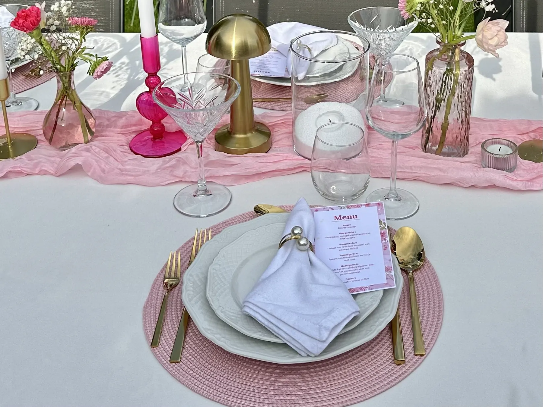 Roze gedekte tafel met een menukaart op het bord, een gouden tafellampje en roze kandelaars