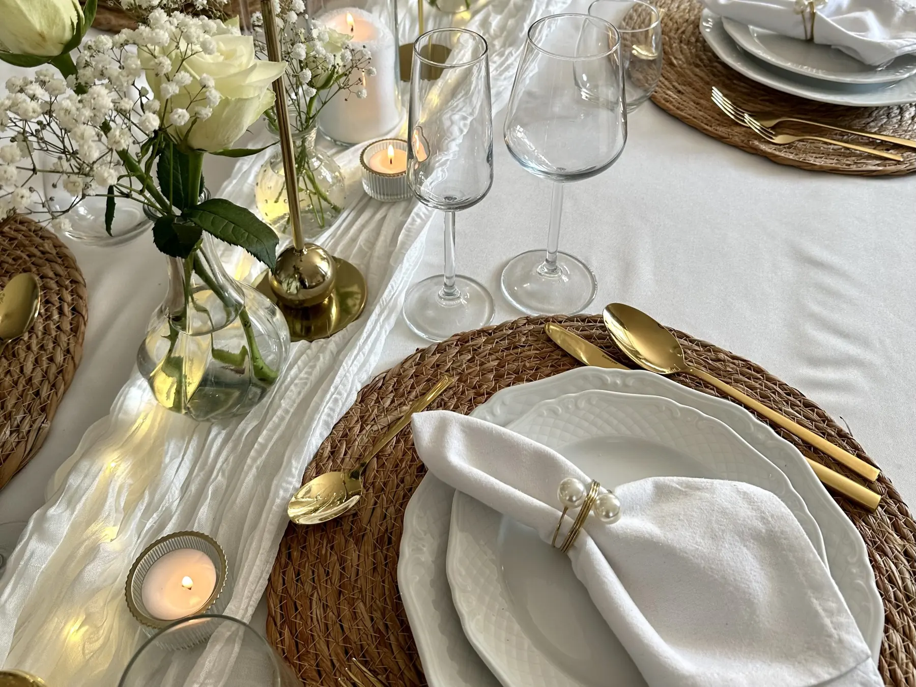 Wit servies op rieten placemats met gouden bestek, witte rozen en waxinelichtjes