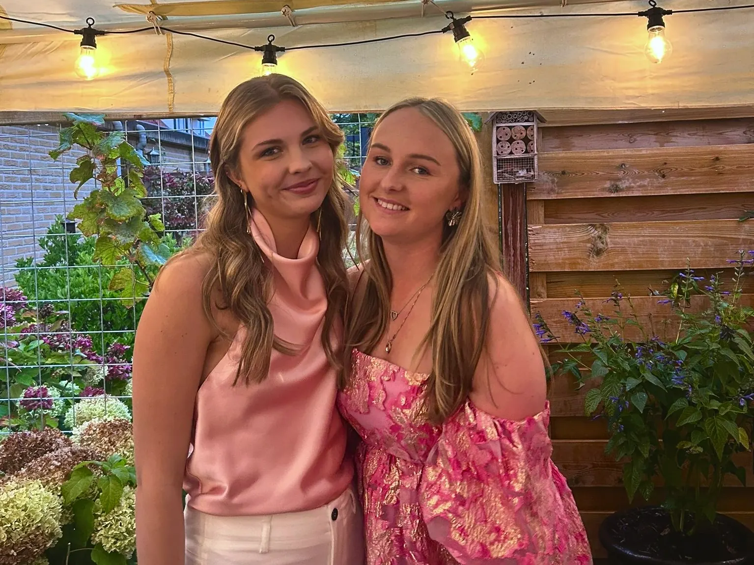 Charlize en Sanne in roze kleding in een tuin met lichtslingers