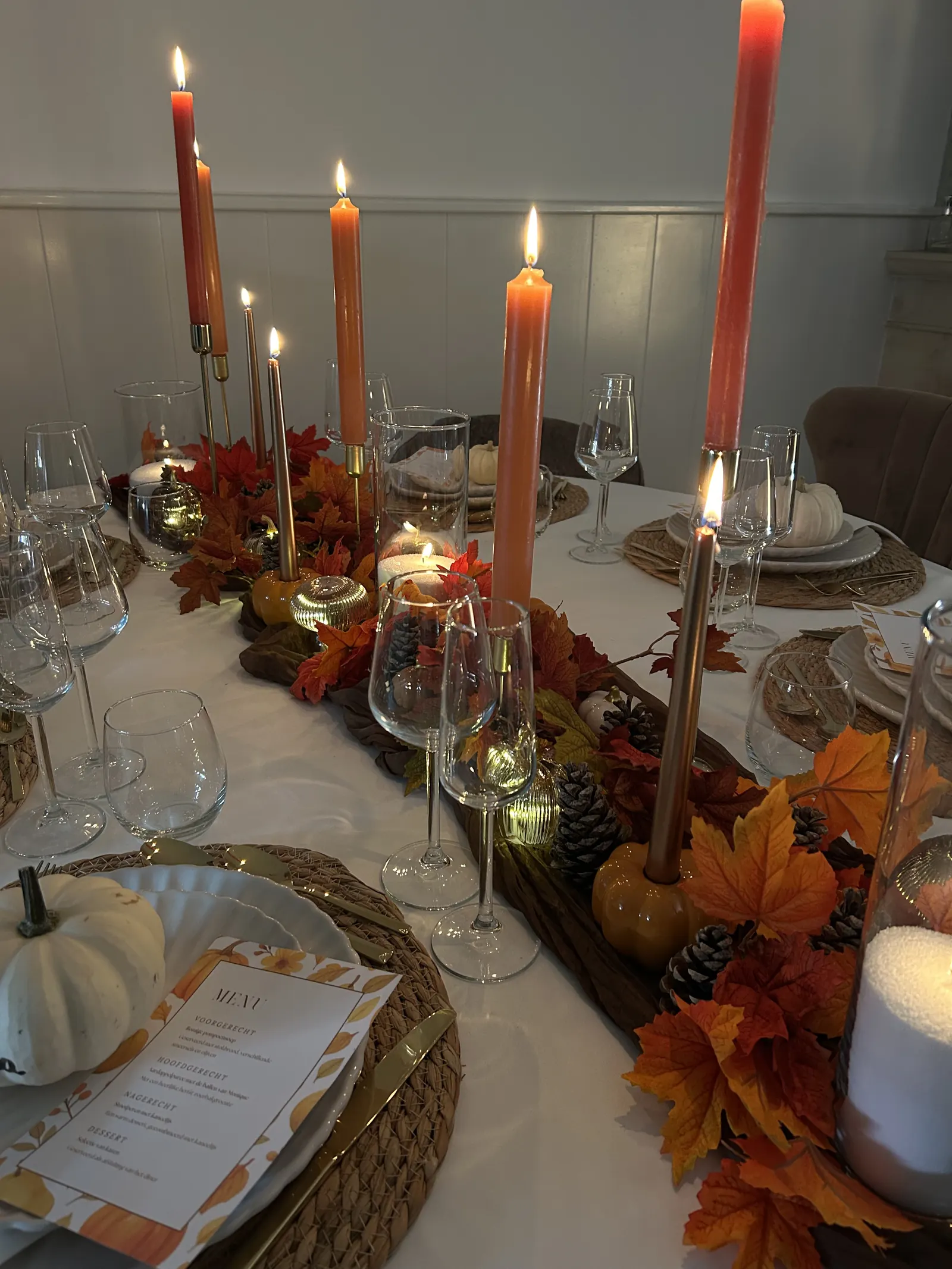 Lange herfsttafel met hoge oranje kaarsen, bladeren en een menukaart bij elk bord