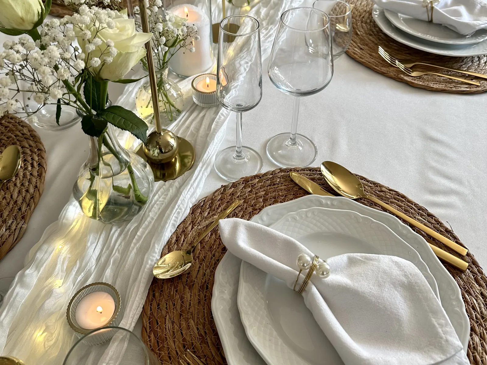 Gedekte tafel met witte rozen, gipskruid, gouden kandelaars en wit servies op rieten placemats