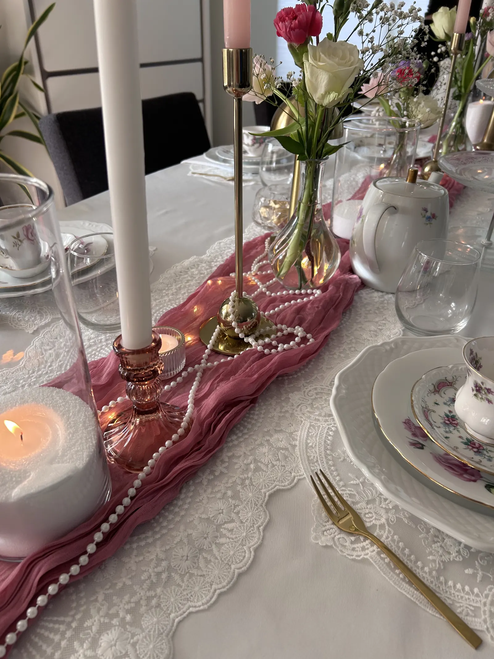 Witte en roze kaarsen, parelkettingen en een theepot op de oudroze tafelloper