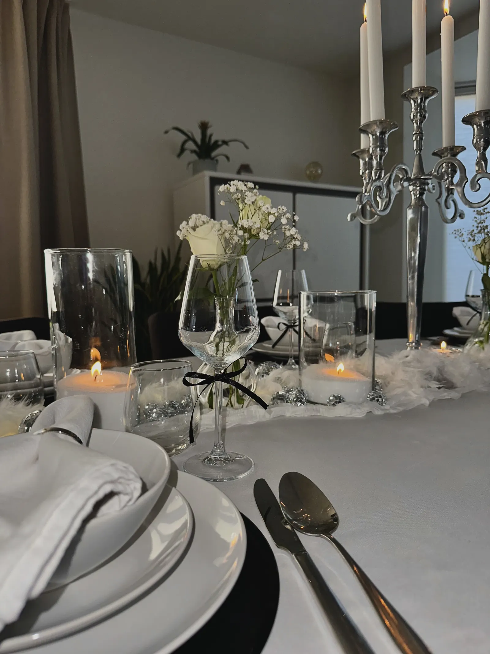 Wijnglas met een zwart strikje bij een bord met zwart onderbord, witte bloemen en kaarslicht