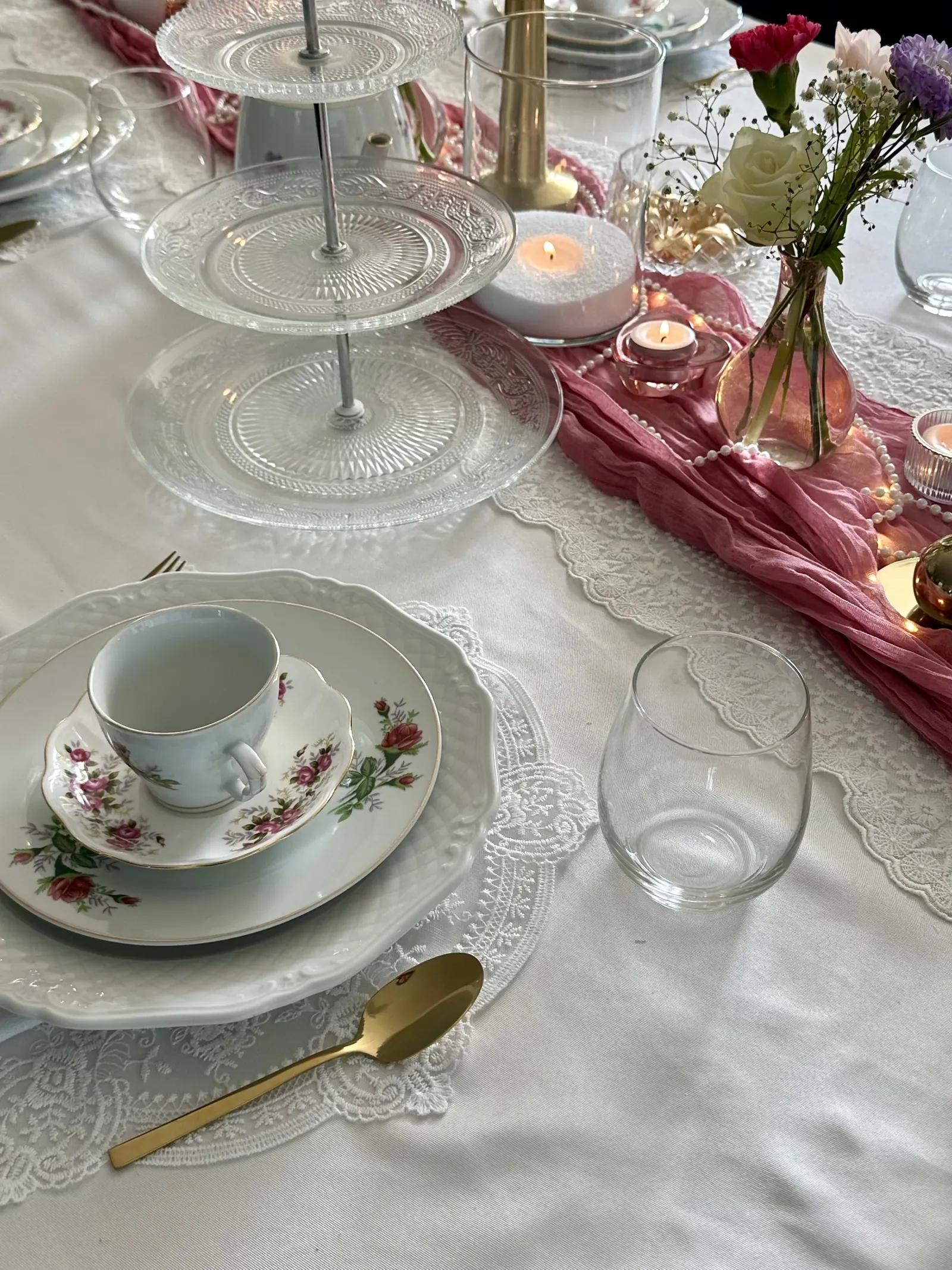 Theekopje en schotel naast een glazen etagère en bloemen op een kanten tafelloper