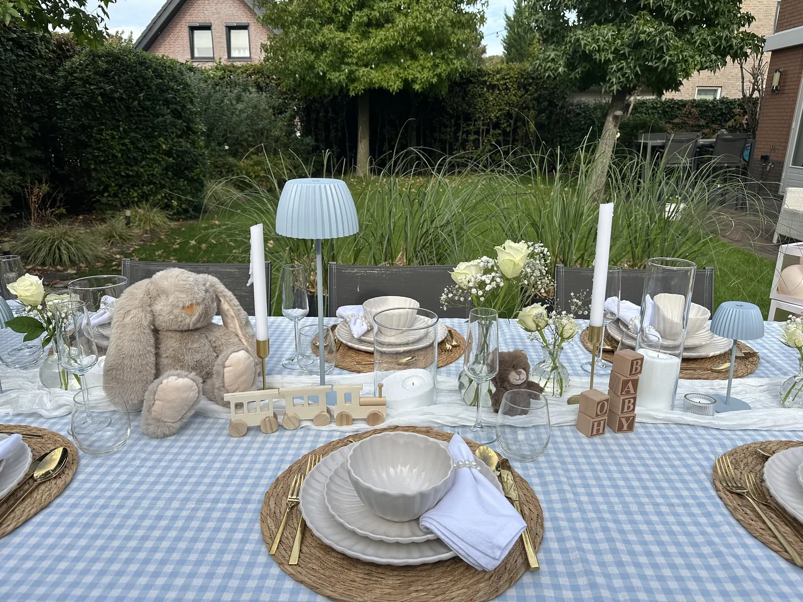 Babyshowertafel in de tuin met een lichtblauw geblokt tafelkleed, knuffelkonijn en houten trein